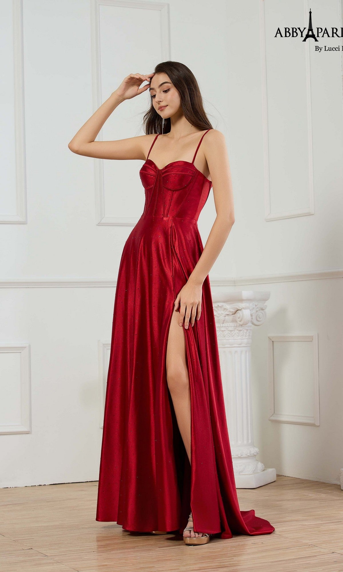 A woman poses indoors in the Abby Paris 90374 long wine prom dress, featuring a sleeveless corset bodice and high slit. The metallic stretch satin fabric catches the light, highlighting her elegant look against a white paneled wall.
